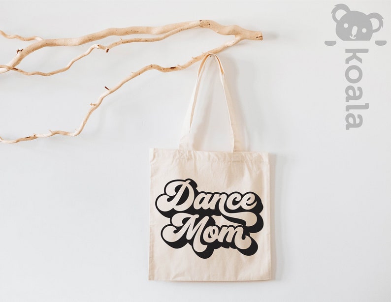以下が含まれることがあります： レトロなフォントで「Dance Mom」と書かれた白いキャンバス地のトートバッグ。白い壁に、流木が後ろに置かれた状態で吊り下げられている。