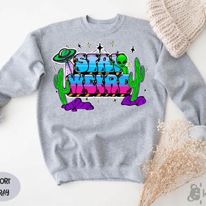 May include: A gray sweatshirt with the text "Stay Weird" in colorful letters, surrounded by stars, a UFO, and cacti.