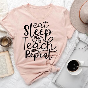 May include: A heather peach colored t-shirt with the text "Eat Sleep Teach Repeat" in black. The text is in a stylized font and includes a graduation cap and pencil graphic.