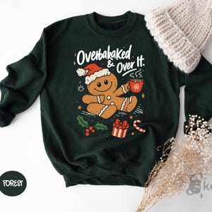 May include: A dark green sweatshirt with a graphic of a gingerbread man wearing a Santa hat and holding a mug. The text "Overbaked & Over It" is above the gingerbread man. A white knit beanie is in the upper right corner.