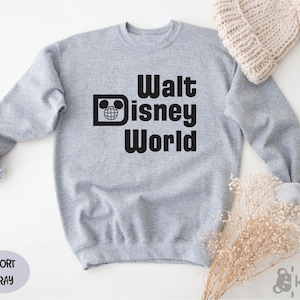 May include: A light gray sweatshirt with the words "Walt Disney World" in bold black letters. The design includes the iconic Mickey Mouse head and a globe. A light gray oval with the words "Sport Gray" is in the lower left corner.