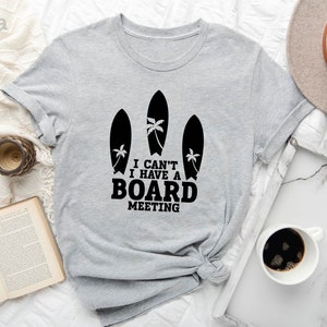 May include: A light gray t-shirt with a graphic of three surfboards and palm trees. The text on the shirt reads "I can't I have a board meeting."