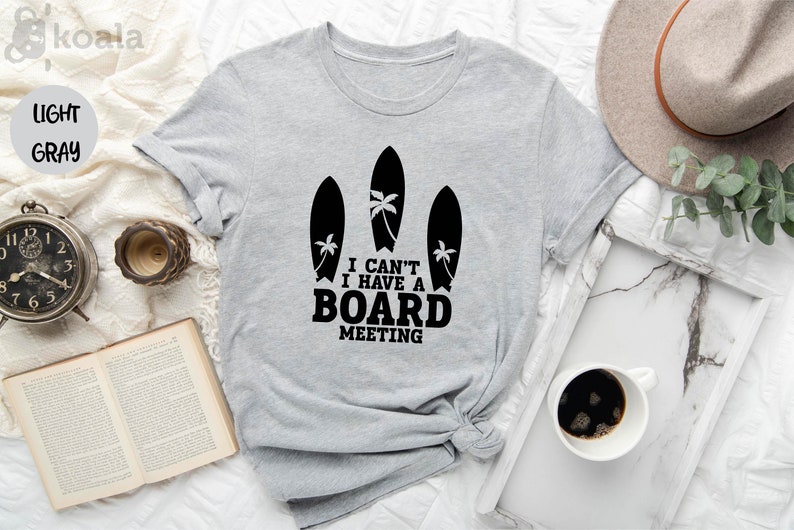 May include: A light gray t-shirt with a graphic of three surfboards and palm trees. The text on the shirt reads "I can't I have a board meeting."