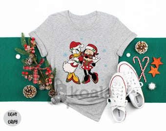 May include: Light gray t-shirt featuring Donald Duck and Minnie Mouse in Christmas attire. The cartoon characters are embracing, surrounded by snowflakes. White sneakers and holiday decorations complete the festive scene.