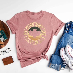 May include: Heather mauve t-shirt with the text "MY OTHER SHIRT HAS CLAY ON IT" in a circular design. The shirt is styled with blue jeans and white sneakers. Other accessories include a wooden tray, camera, and glasses.