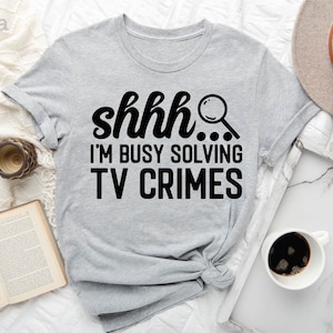 May include: A light gray t-shirt with the text "shhh... I'm busy solving TV crimes" in black. A magnifying glass icon is next to the text.