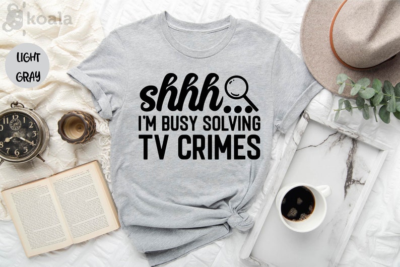 May include: A light gray t-shirt with the text "shhh... I'm busy solving TV crimes" in black. A magnifying glass icon is next to the text.