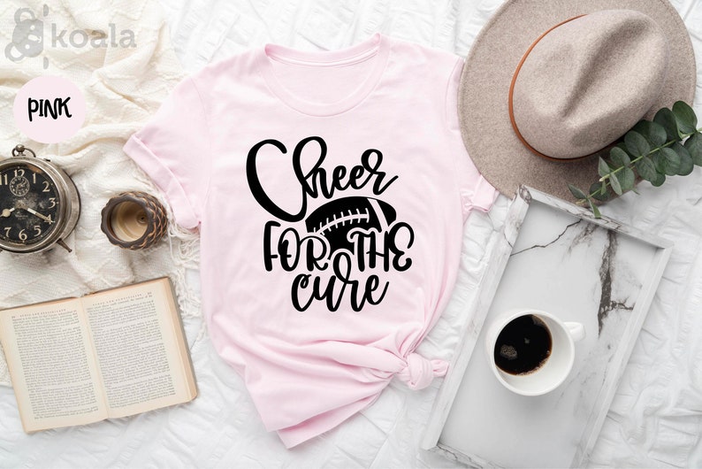 May include: Pink t-shirt with black text that reads "Cheer for the cure" and a black football graphic.