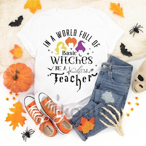 May include: White t-shirt with the text "IN A WORLD FULL OF BASIC WITCHES BE A TEACHER" in black. The shirt is paired with blue jeans, orange sneakers, and Halloween decorations like a pumpkin, leaves, and a skull.