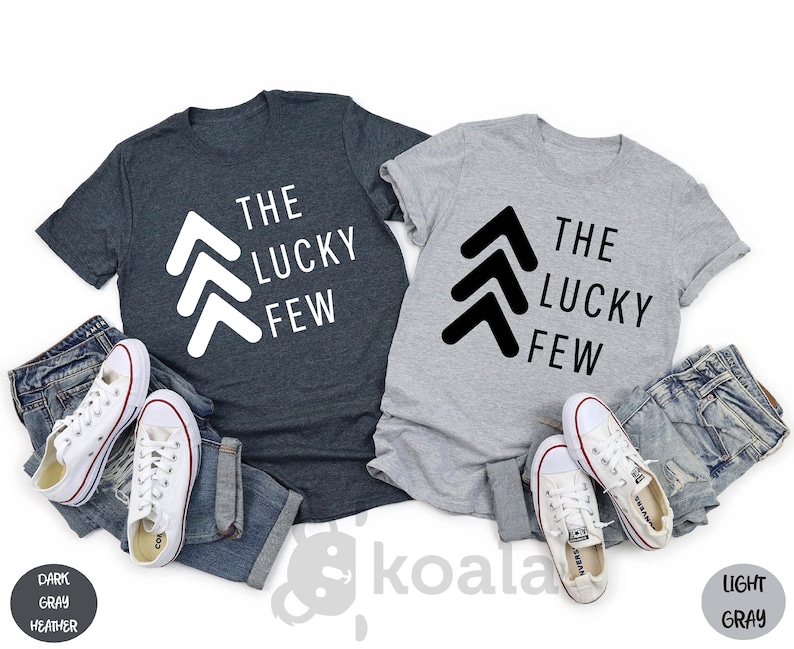May include: Two t-shirts, one dark heather gray and one light gray, with the text "The Lucky Few" and a graphic of three white arrows pointing upwards on the dark heather gray shirt and three black arrows pointing upwards on the light gray shirt.