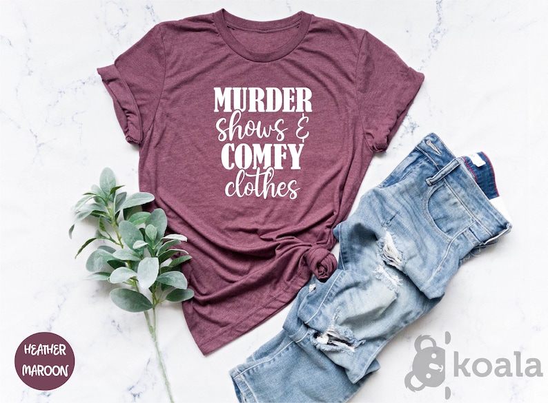 May include: A maroon t-shirt with the text "MURDER shows & COMFY clothes" in white. The shirt is styled with distressed blue jeans. A sprig of green leaves is placed near the shirt and jeans.
