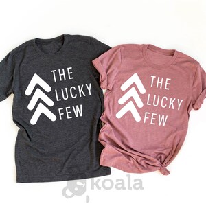 May include: Two t-shirts with the text "The Lucky Few" and three white arrows pointing upwards. The shirt on the left is dark heather gray and the shirt on the right is heather mauve.