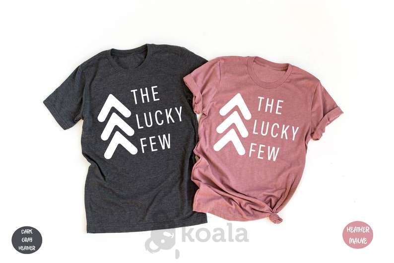 May include: Two t-shirts with the text "The Lucky Few" and three white arrows pointing upwards. The shirt on the left is dark heather gray and the shirt on the right is heather mauve.