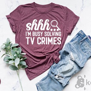 May include: A heather maroon t-shirt with the text "shhh... I'm busy solving TV crimes" in white lettering. The shirt is shown on a white marble surface with a sprig of greenery and a pair of blue jeans.