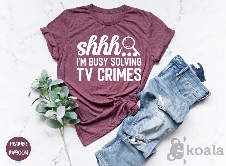 May include: A heather maroon t-shirt with the text "shhh... I'm busy solving TV crimes" in white lettering. The shirt is shown on a white marble surface with a sprig of greenery and a pair of blue jeans.