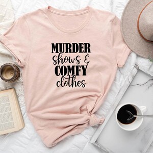 May include: A pale peach t-shirt with the words "MURDER shows & COMFY clothes" printed in black. The shirt is displayed on a white surface with a book, a clock, a hat, and a coffee cup.