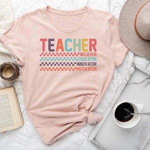 May include: A heather peach t-shirt with the word "TEACHER" in rainbow colors and the words "BELIEVER", "EDUCATOR", "INNOVATOR", and "MOTIVATOR" in smaller text below.