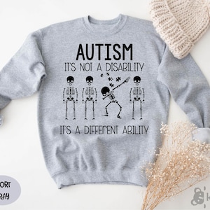 May include: Gray sweatshirt with the text "AUTISM IT'S NOT A DISABILITY IT'S A DIFFERENT ABILITY" and a skeleton doing a dab with puzzle pieces around it. There are three other skeletons standing in a line.