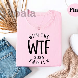 a pink t-shirt with the text "WTF 2026 FAMILY" printed on it, along with a pair of jeans and a pair of white sneakers. The t-shirt is laid out on a white background, and there are some decorative elements such as a palm leaf and a beige blanket nearby.