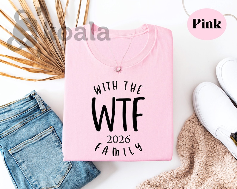 a pink t-shirt with the text "WTF 2026 FAMILY" printed on it, along with a pair of jeans and a pair of white sneakers. The t-shirt is laid out on a white background, and there are some decorative elements such as a palm leaf and a beige blanket nearby.