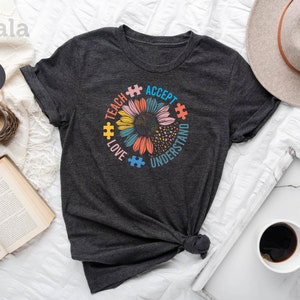 May include: Dark gray heather t-shirt with a colorful sunflower design and the text "Teach Love Accept Understand" around the flower. The sunflower is made up of puzzle pieces.