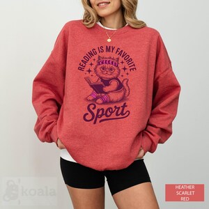 A woman wearing a red sweatshirt with a graphic design of a cat reading a book and the text "Reading is my favorite Sport" printed on it.