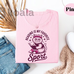 a pink t-shirt with a cat graphic and the text "Reading is my favorite sport" printed on it, along with a pair of jeans and a pair of white sneakers.