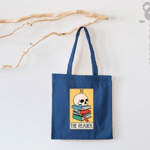 May include: A blue tote bag with a tarot card design featuring a skull on top of a stack of books. The card reads "XII THE READER."