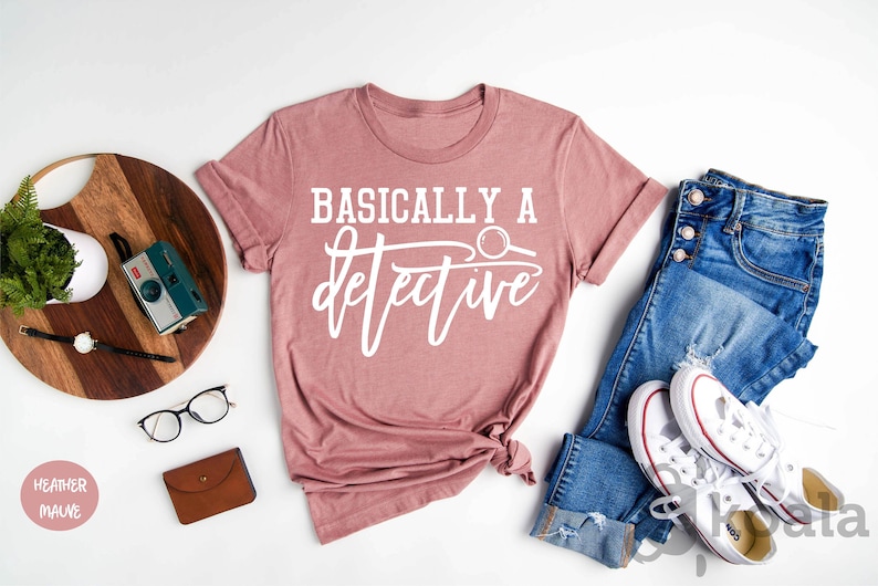 May include: A heather mauve t-shirt with the text "Basically a detective" in white lettering. The shirt is folded and laid flat on a white surface with a pair of glasses, a wooden cutting board, a small potted plant, a vintage camera, a brown wallet, and a pair of blue jeans with white sneakers.