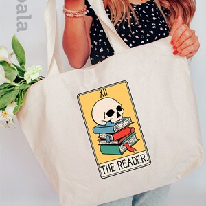 May include: A white canvas tote bag with a zipper closure. The bag features a graphic of a tarot card, The Reader, with a skull and books. The bag is 20 inches wide and 15 inches high and is made of 100% cotton.