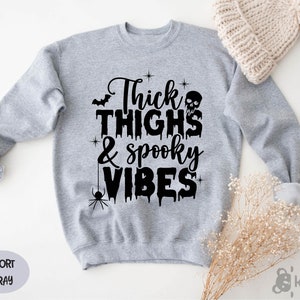 Op de afbeelding: Een lichtgrijs sweatshirt met de zwarte tekst "Thick Thighs & Spooky Vibes". Het ontwerp bevat een vleermuis, een schedel, een spin en sterren. Het sweatshirt is sportgrijs van kleur.