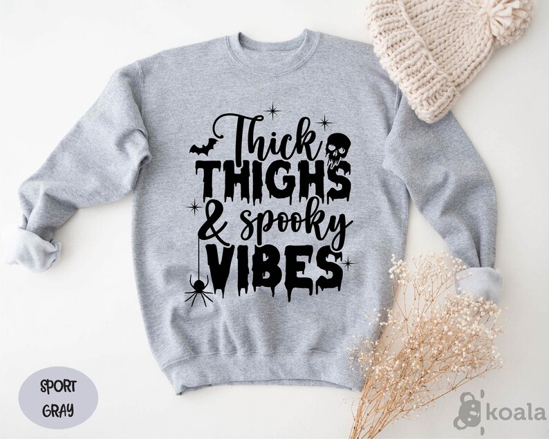 Op de afbeelding: Een lichtgrijs sweatshirt met de zwarte tekst "Thick Thighs & Spooky Vibes". Het ontwerp bevat een vleermuis, een schedel, een spin en sterren. Het sweatshirt is sportgrijs van kleur.