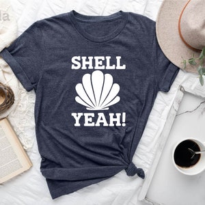 May include: A heather navy blue t-shirt with the text "SHELL YEAH!" in white, featuring a white seashell graphic.