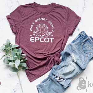 May include: A heather maroon t-shirt with a white graphic design featuring the text "Walt Disney World 1971 1982 EPCOT" and a white outline of the Spaceship Earth icon.
