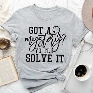 Puede incluir: Camiseta gris claro con texto negro que dice "GOT A mystery? YO, I'LL SOLVE IT" y un gr&aacute;fico de lupa negra.