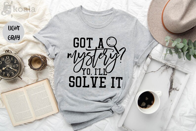 Puede incluir: Camiseta gris claro con texto negro que dice "GOT A mystery? YO, I'LL SOLVE IT" y un gr&aacute;fico de lupa negra.