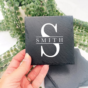 May include: Black slate coasters with a white monogram "S" and the name "SMITH" printed below. The coasters are square and have a natural, textured surface. The background includes greenery.