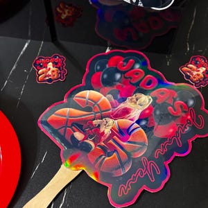 May include: A holographic fan with a basketball theme. The fan features a basketball player and the text "Jordan Year" in a cursive font. The fan is attached to a wooden stick. Additional basketball-themed decorations are scattered around.