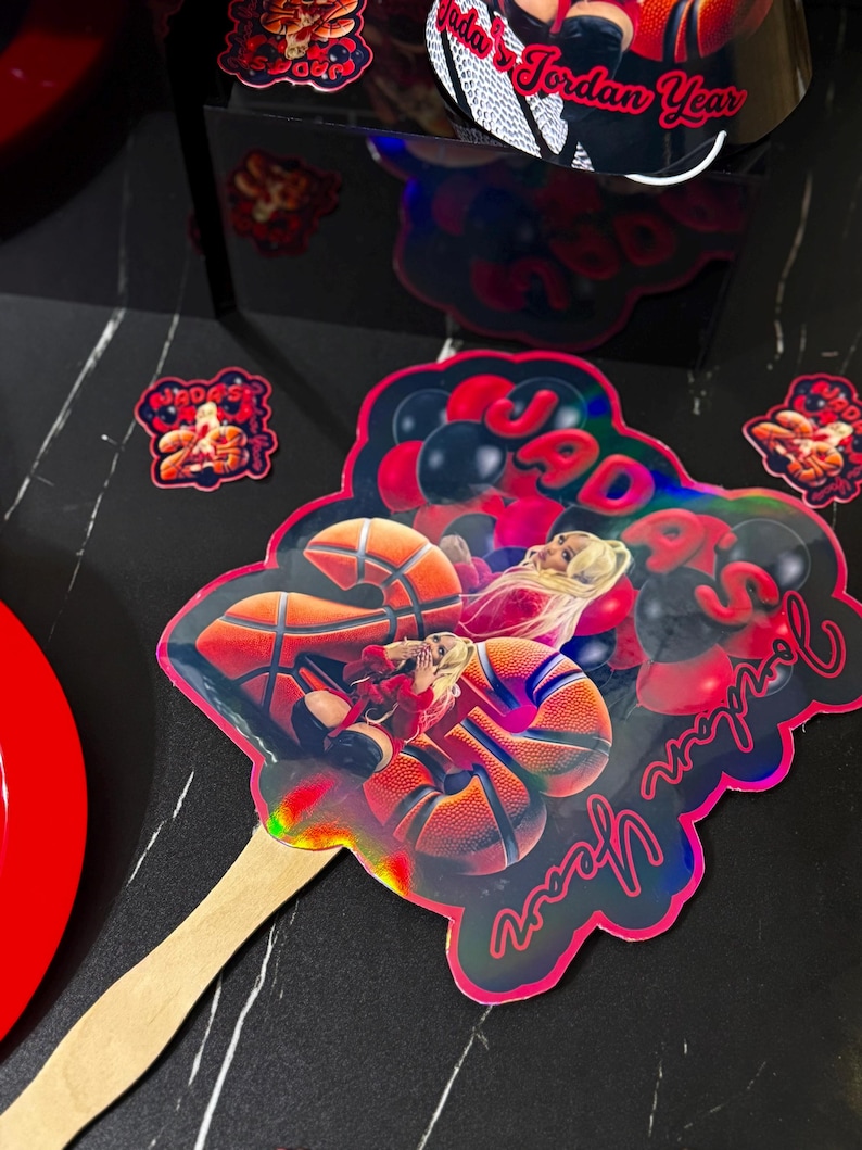 May include: A holographic fan with a basketball theme. The fan features a basketball player and the text "Jordan Year" in a cursive font. The fan is attached to a wooden stick. Additional basketball-themed decorations are scattered around.