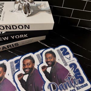 May include: A stack of books with the titles "LONDON", "NEW YORK", and "PARIS" is topped with a silver balloon dog sculpture. Several fan-shaped photo props with a man's image and text that reads "CONGRATS 25 Dante United States Airman" are in the foreground.