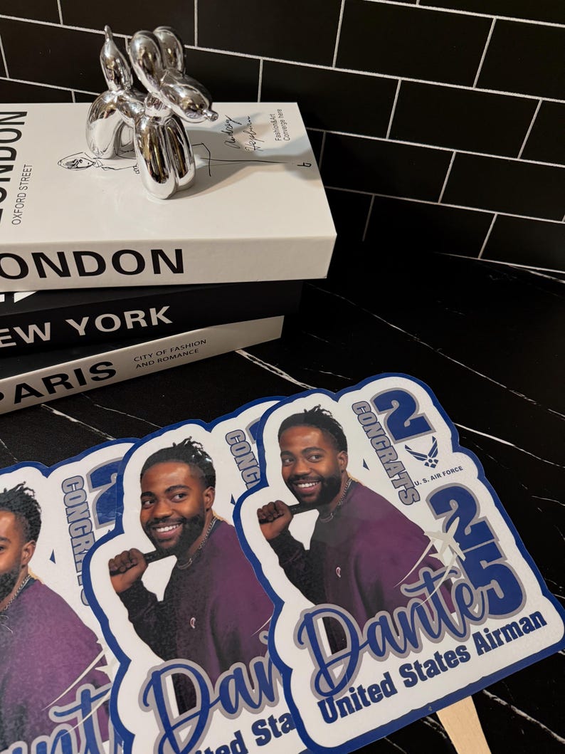 May include: A stack of books with the titles "LONDON", "NEW YORK", and "PARIS" is topped with a silver balloon dog sculpture. Several fan-shaped photo props with a man's image and text that reads "CONGRATS 25 Dante United States Airman" are in the foreground.