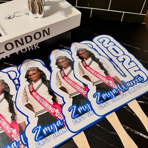 May include: Several hand fans with a photo of a young woman wearing a sash that reads "2024 Homecoming Queen." The fans have a blue and white design with the text "Z'mya Warren" and "East Orange Campus High."
