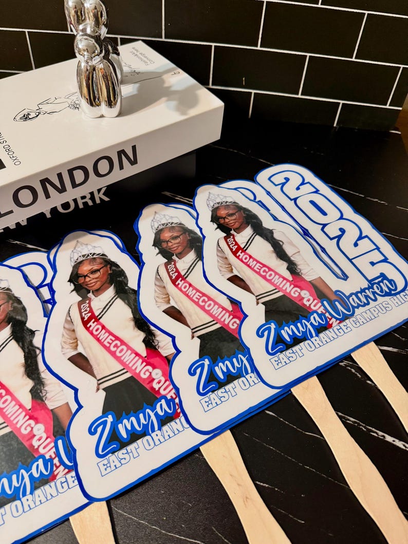 May include: Several hand fans with a photo of a young woman wearing a sash that reads "2024 Homecoming Queen." The fans have a blue and white design with the text "Z'mya Warren" and "East Orange Campus High."