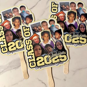 May include: Three graduation fans with a collage of photos, the text "CLASS OF 2023", and the name "Jahira". The fans have a yellow and black design with a wooden handle. The fans are on a white marble surface.