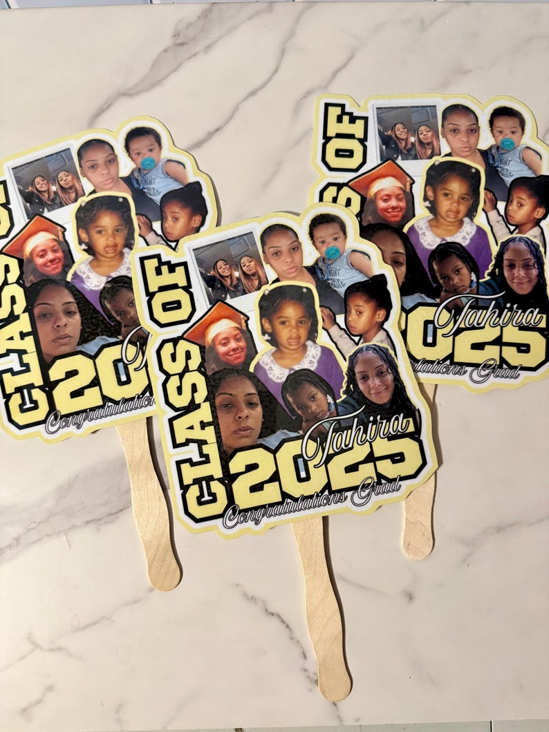 May include: Three graduation fans with a collage of photos, the text "CLASS OF 2023", and the name "Jahira". The fans have a yellow and black design with a wooden handle. The fans are on a white marble surface.