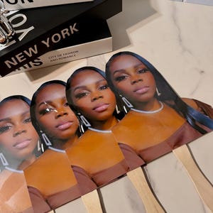 May include: A collection of four fan-shaped face cards featuring a woman with long dark hair, wearing a brown top and silver jewelry. The cards are arranged in a row, with wooden sticks attached to the bottom. Decorative books are in the background.