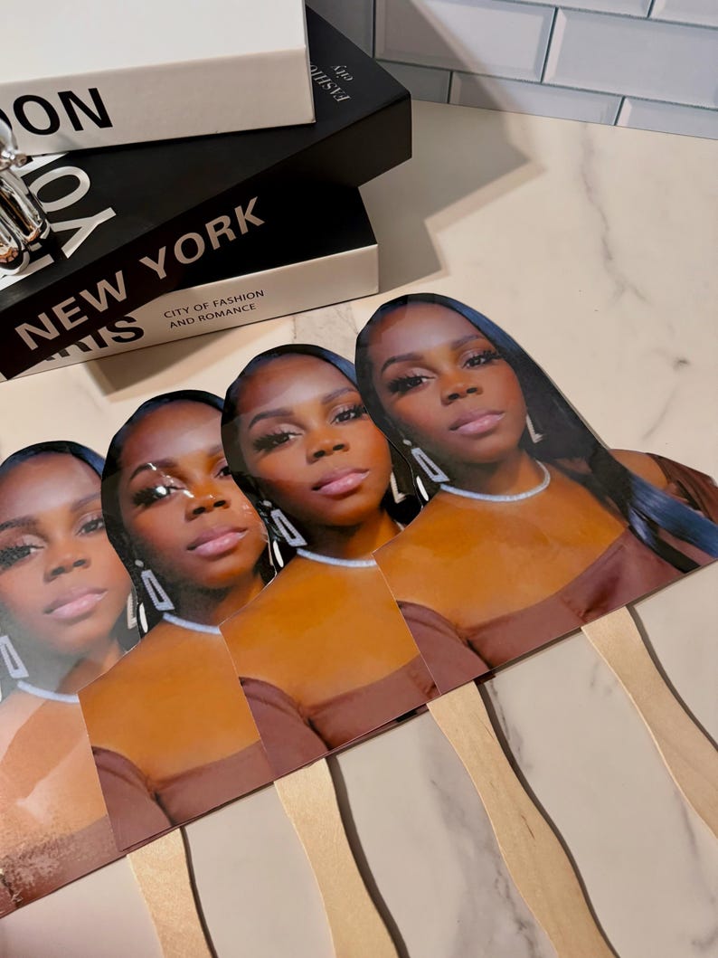 May include: A collection of four fan-shaped face cards featuring a woman with long dark hair, wearing a brown top and silver jewelry. The cards are arranged in a row, with wooden sticks attached to the bottom. Decorative books are in the background.