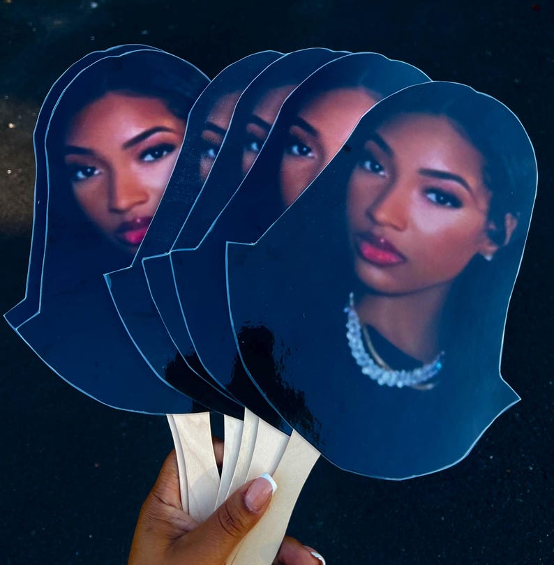 May include: A collection of five dark blue fans, each featuring a portrait of a woman with dark hair, red lipstick, and a necklace. The fans are held together by wooden sticks.