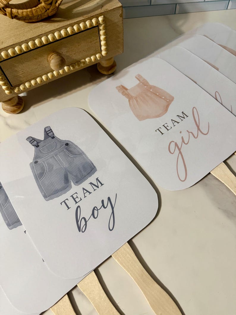 May include: A collection of gender reveal fans. One fan features a watercolor illustration of blue overalls with the text "TEAM BOY." Another fan displays a pink romper with the text "TEAM girl." The fans have wooden handles.