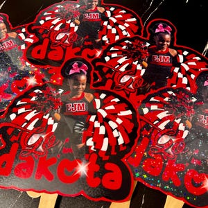 May include: A collection of red, black, and white cheerleading-themed cutouts. Each cutout features a young girl in a cheerleading uniform with the text "Go Dakota". The cutouts have wooden sticks attached.